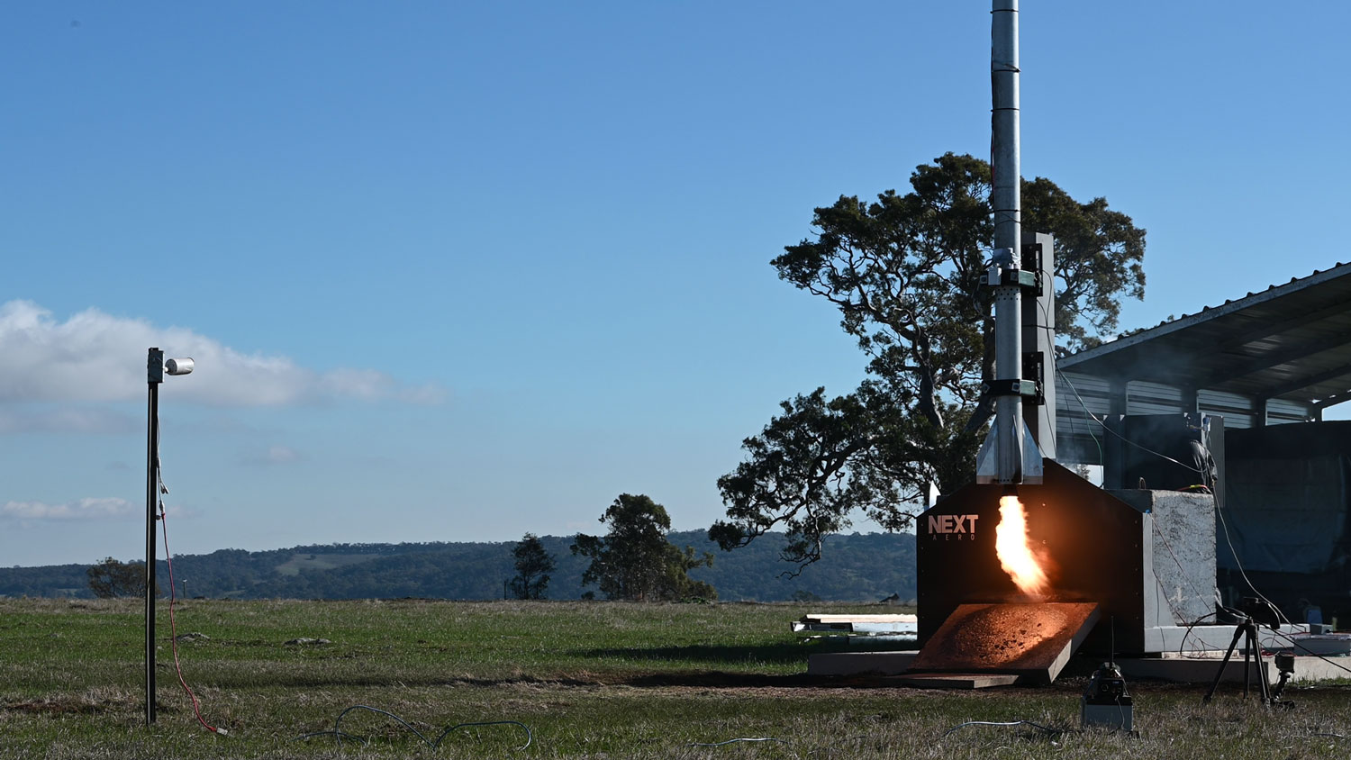 To the right of a grassy outdoor landscape with a blue sky, a rocket is set up on a testing platform with a flame emerging from underneath.