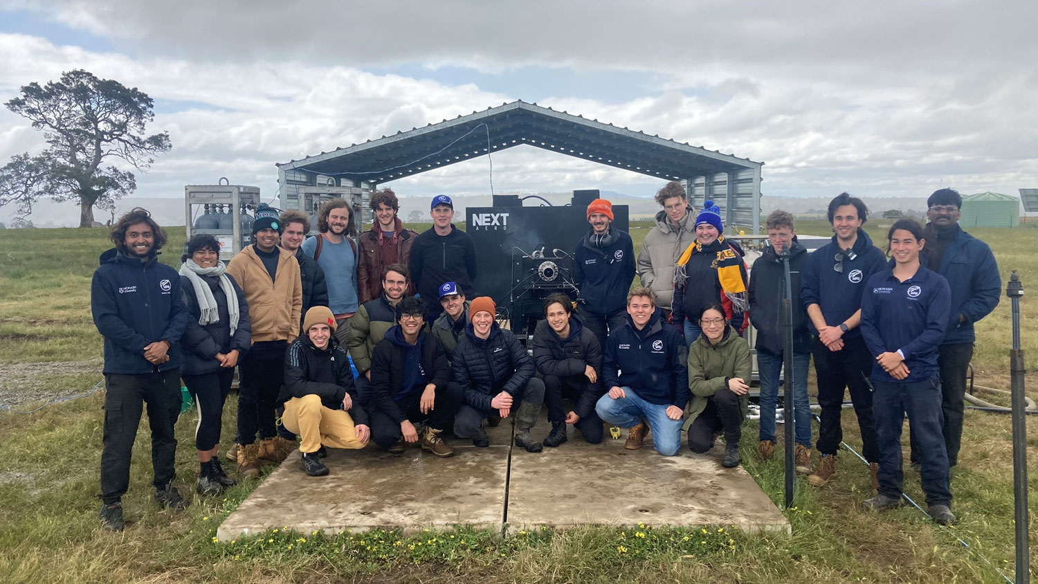 A large group of Next Aero staff pose together for a picture outdoors, with their engine testing platform behind them.