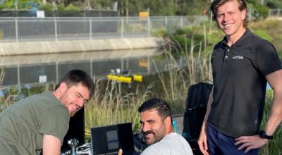 A group of three men with equipment by a riverbank. The one of the left is crouching, the middle one is sitting and the one on the right is standing. All look relatively young