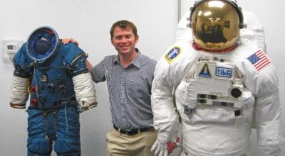 A white middle aged man standing between two spacesuits. A blue one on his right and a white one on his left facing the camera