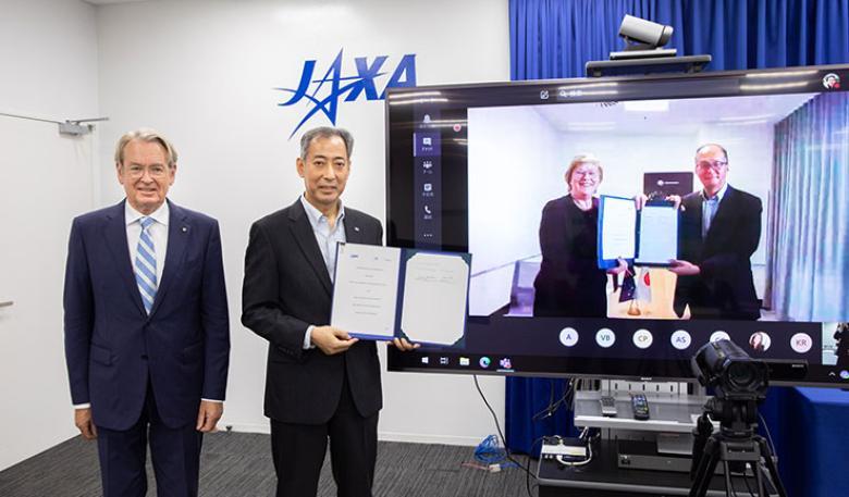 Presentation of signed agreement between Australia and Japan. Two men standing beside a tv screen, with two people featured on the screen