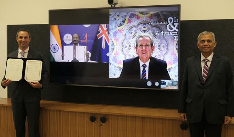 Two men standing in front of a television, with a third man on the screen. The man on the left holds the signed documents.