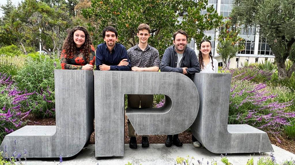 Five people in casual clothes stand behind a large 'JPL' stone monument on the ground in a garden area.