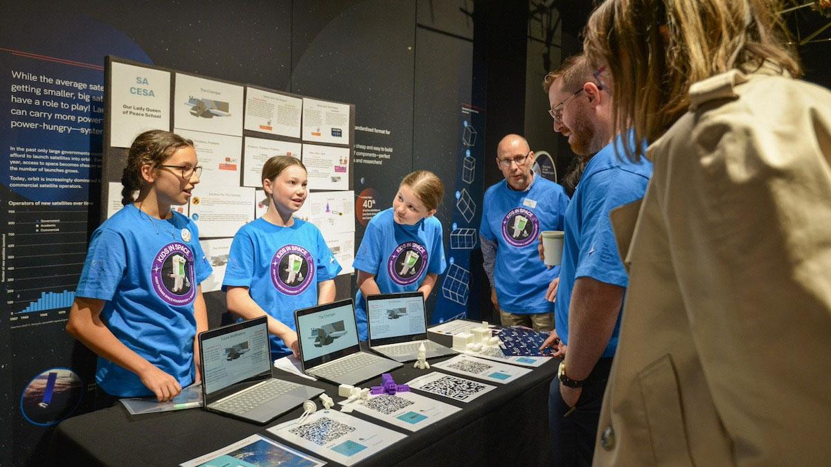 Our Lady Queen Of Peace students in blue shirts stand behind the table displaying their design, as they present to two people in front of the table.