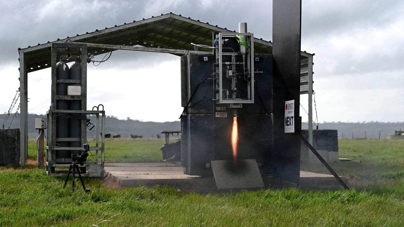 In the distance in an outdoor landscape surrounded by grass, an engine is set up on a platform and tested with a flame emerging from underneath.