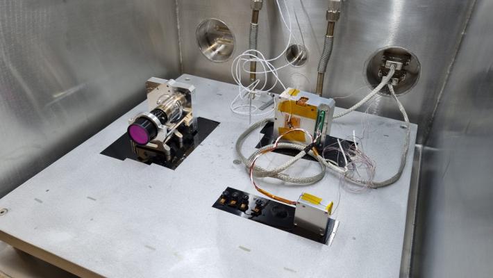 Three separate components sit on a flat surface inside a box-shaped testing apparatus with silver walls, with various wires connected to them.