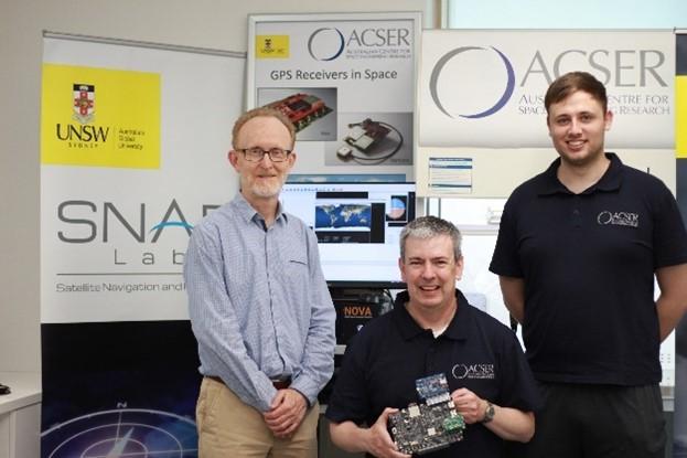 The ACSER Team of three standing in front of a computer and various signs. The middle person is holding the GNSS prototype