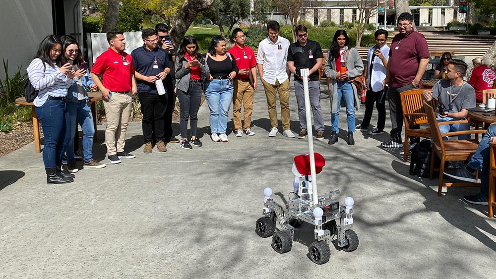 A crowd of people in the background watch Akin robot Henry The Helper in the foreground, during a demonstration in a courtyard at NASA JPL